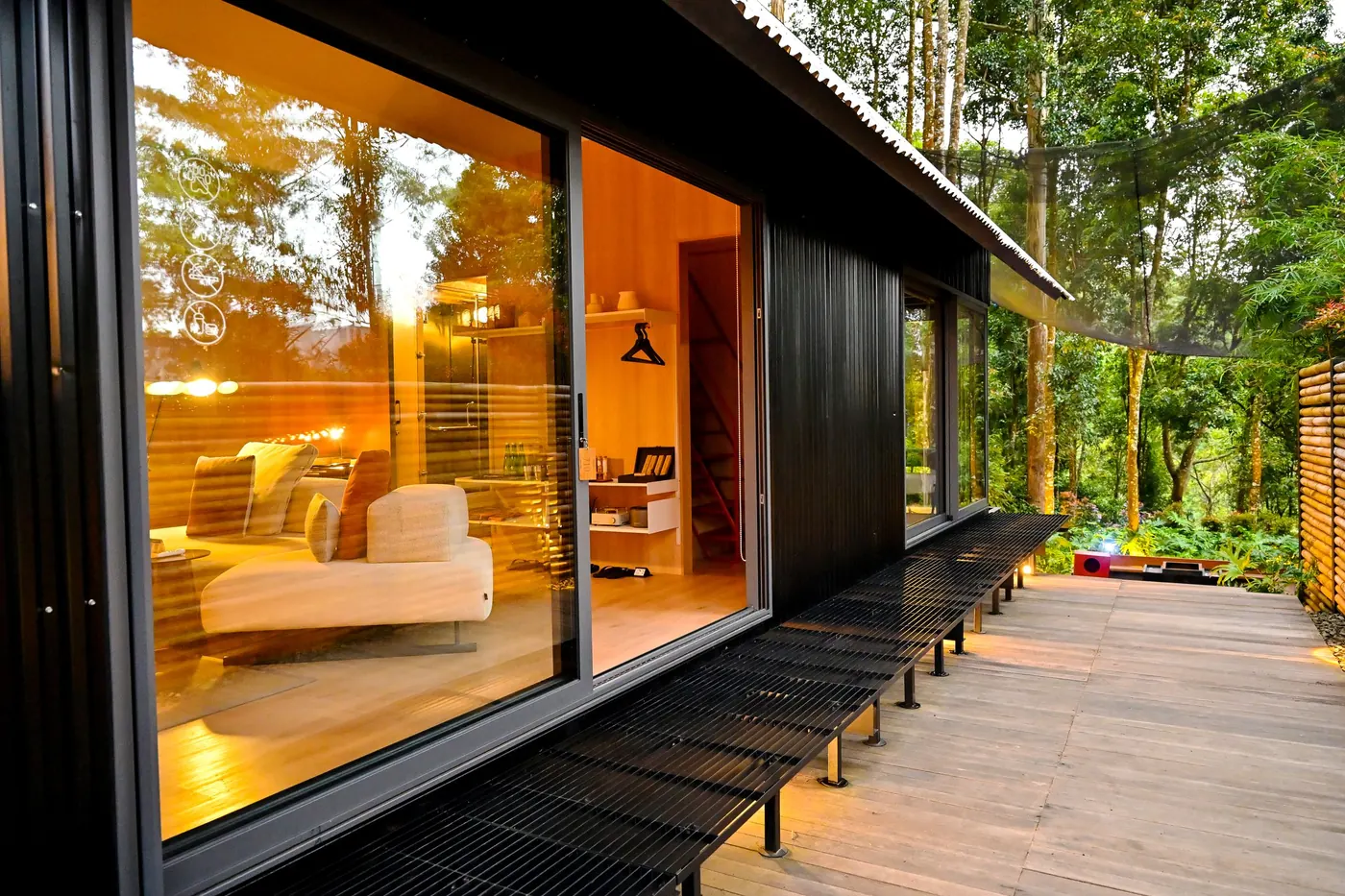 A dark timber cabin at dusk with full-height glass doors, the living room lit warm inside, and a wide deck along the front.