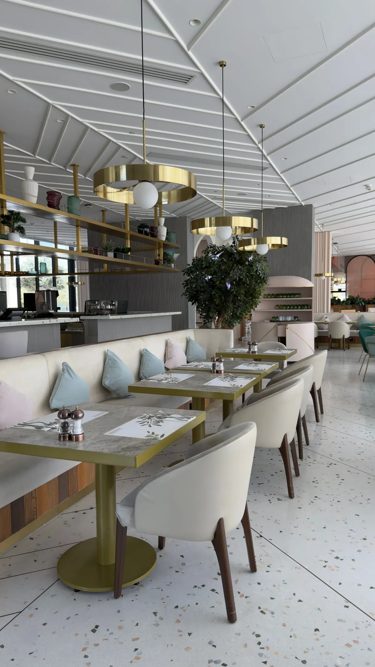A bright restaurant with a terrazzo floor, a long banquette with pale blue cushions, cream chairs and brass shelving under a white ribbed ceiling.