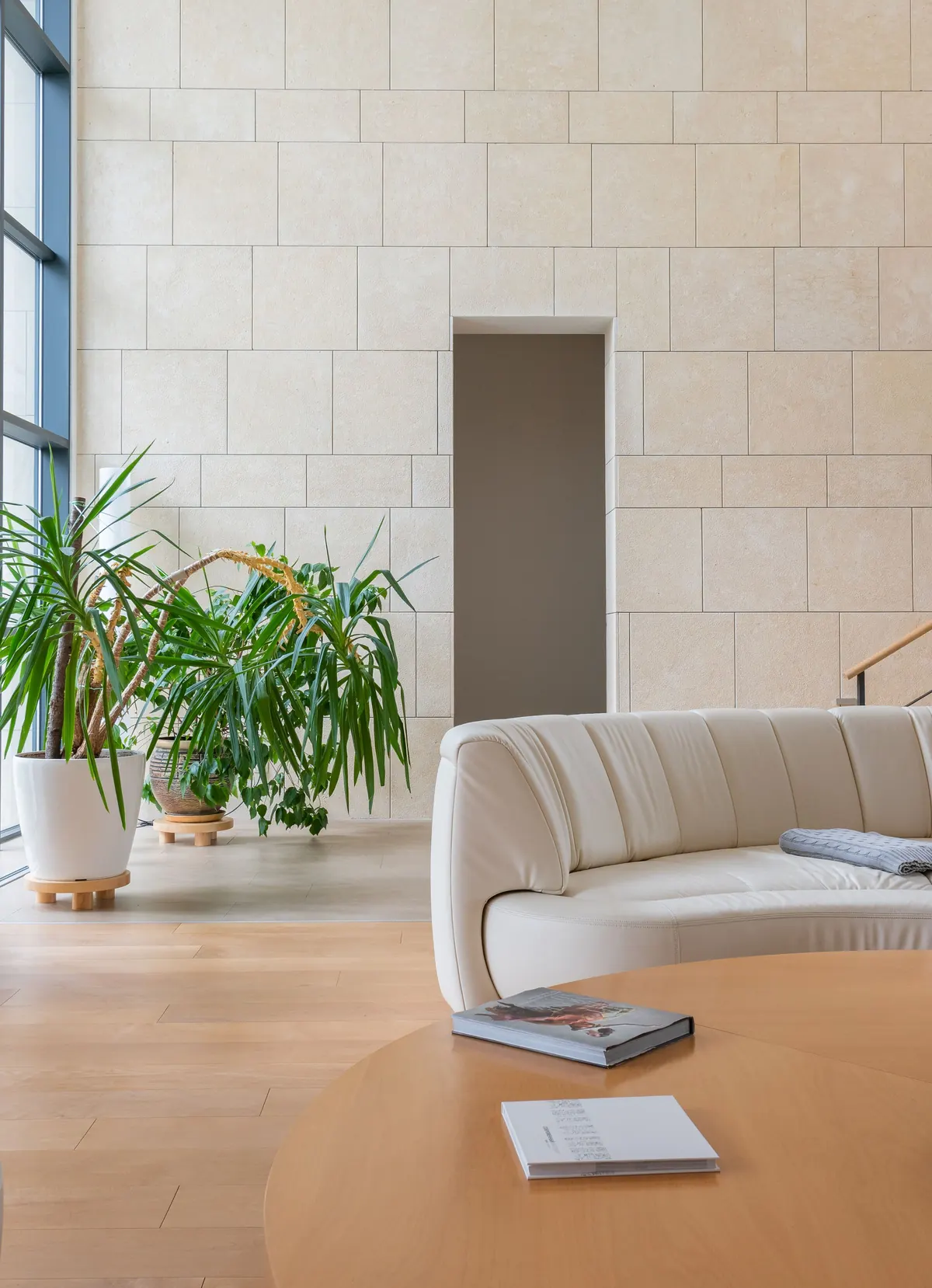 A curved cream sofa and a round oak table in front of a pale limestone wall, with tall plants by the window.