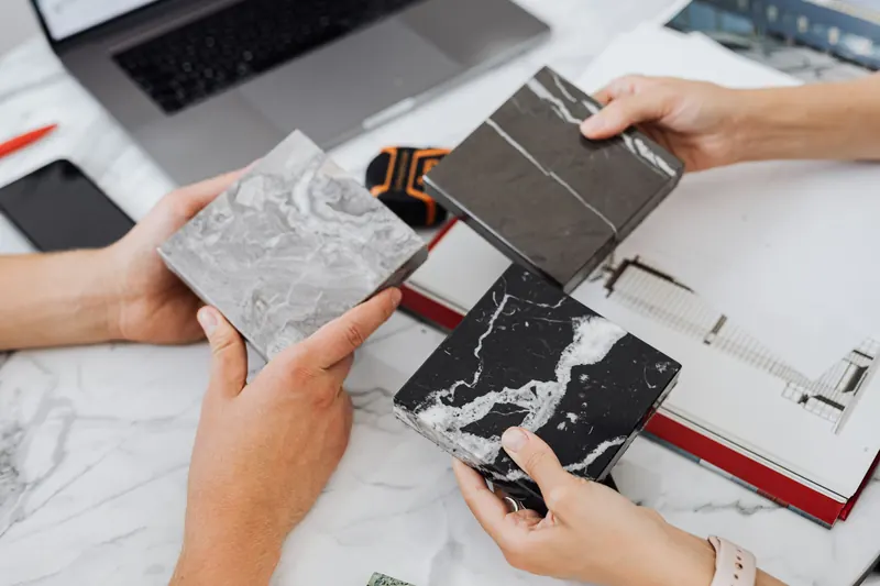 Two pairs of hands comparing marble tile samples over drawings on a desk.